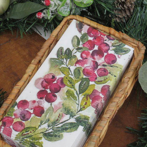 Rattan napkin holder with a stack of white paper napkins featuring a watercolor-style cranberry and greenery design. Displayed on a wooden surface with frosted greenery and a pinecone in the background.  