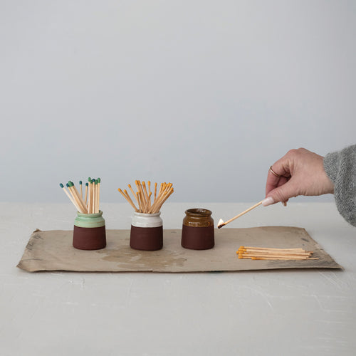 Three ceramic matchstick holders with strikers in green, white, and brown. Person striking a match on brown holder. Holders on a tray against a white background. 