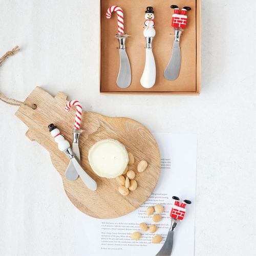 Set of three holiday cheese spreaders with glass handles shaped like a candy cane, a snowman, and Santa's legs in a chimney, displayed on a snowman shaped wooden board with nuts and cheese beside a set of matching spreaders in a brown packaging box. Against a white background. 