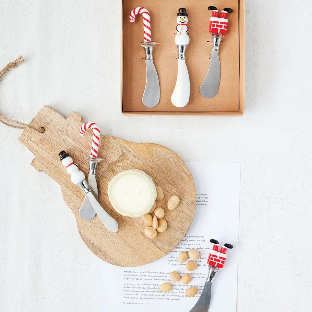 Set of three holiday cheese spreaders with glass handles shaped like a candy cane, a snowman, and Santa's legs in a chimney, displayed on a snowman shaped wooden board with nuts and cheese beside a set of matching spreaders in a brown packaging box. Against a white background. 