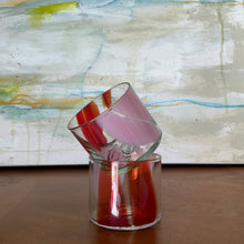 Load image into Gallery viewer, Splash Cups clear hand-blown glass set of two with abstract light pink and red swirled design, stacked on one another on  wooden surface against abstract background.
