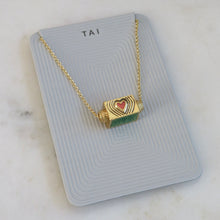 Load image into Gallery viewer, Gold-plated rectangular spinner pendant necklace with a red heart motif and radiating line design, displayed on a light gray TAI jewelry card on a white marble background. 
