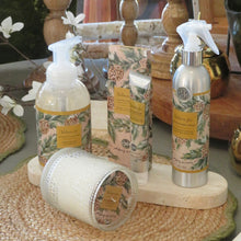 Load image into Gallery viewer, Balsam &amp; Fir, lifestyle image of a soy candle in textured clear glass lying on its side, foaming hand wash, boxed hand cream, and room and fabric spray featuring coordinating cream backgrounds, green evergreen branches, brown pinecones, and gold-colored panels with the scent name and product type in dark lettering, arranged on cream stone trays and a brown woven mat with green scalloped edging.