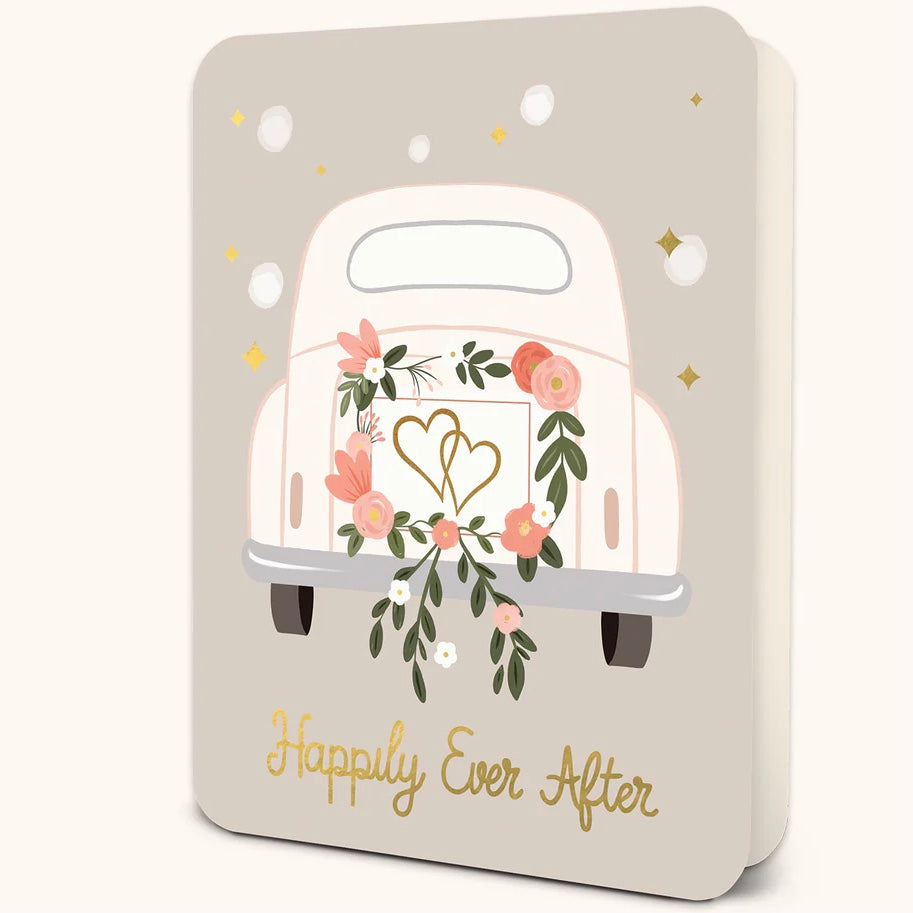 Slightly opened standing pale gray greeting card with white polka dots and gold stars.  In the middle of the card is the back end of white car. Car has a pink and green floral wreath with intertwined hearts on it.. 