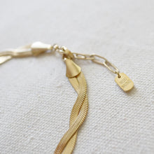 Load image into Gallery viewer, Close-up of an IP gold-plated stainless steel bracelet showcasing the intertwined twisted snake-chain strands, extender chain, and a small "WEST NATIVE" label charm, displayed on a soft white textured surface. 