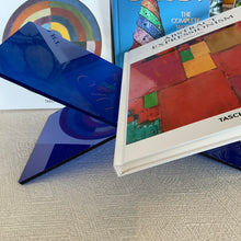 Load image into Gallery viewer, Blue Acrylic Book Holder, transparent blue acrylic book holder with interlocking X-shaped design holding a hardcover book, displayed on beige textured surface with books in the background. 