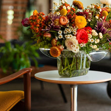 Load image into Gallery viewer, Clear cowboy hat-shaped bowl used as a vase holding a colorful floral arrangement on a small round white table. 