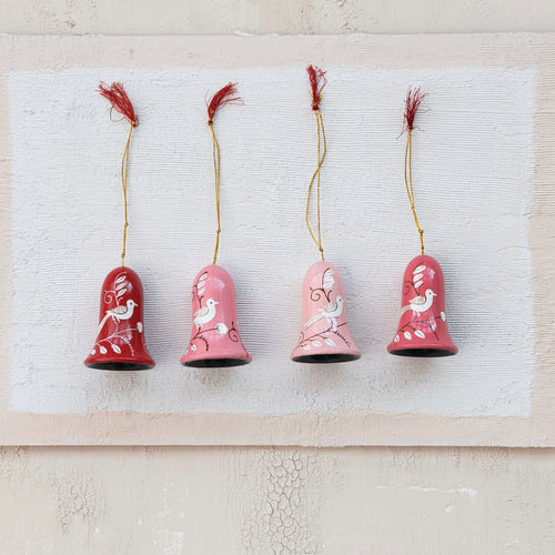 Four wooden bell ornaments in red, pink, light pink, and salmon with a white bird and botanical design, each topped with a gold hanging cord and a red tassel, displayed against a textured cream background. 