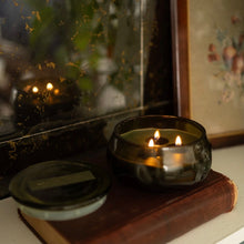 Load image into Gallery viewer, Green lit candle in a green glass container,  with lid beside it. Candle and lid sitting on a worn brown book , on top of a white surface. Black marble looking background and a framed floral picture.