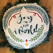 Load image into Gallery viewer, Close-up on a white round ornament-shaped pillow embroidered with &quot;Joy To The World&quot; in green lettering surrounded by green pine branches, red ribbon accents, and yellow stars, displayed on a wooden surface with green garland in the background.