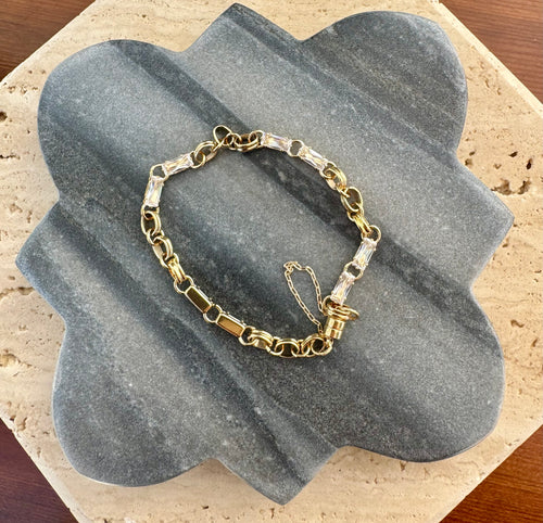 Gold link bracelet accented with square-cut clear crystals, displayed in a loose circle on a quatrefoil-shaped gray stone tile layered over a beige travertine slab on a wooden surface.  