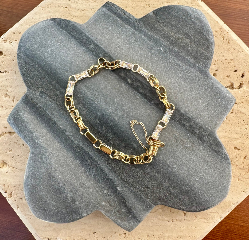 Gold link bracelet accented with square-cut clear crystals, displayed in a loose circle on a quatrefoil-shaped gray stone tile layered over a beige travertine slab on a wooden surface.  