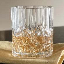 Load image into Gallery viewer, Clear carved lowball tumbler with a faceted pattern holding a light-colored beverage on a beige folded cloth. 