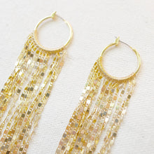 Load image into Gallery viewer, Pair of IP gold-plated hoop earrings with multiple shimmering faceted gold chain strands draped from rounded hoops, displayed on a soft white textured surface. 
