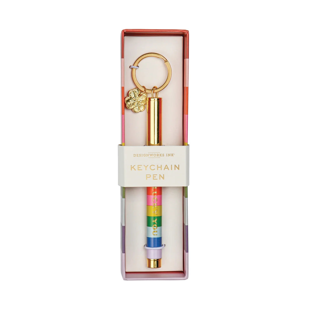 Box on a white background. Inside the background is rainbow striped pen on a gold keychain. The keyring also has a shamrock charm. 