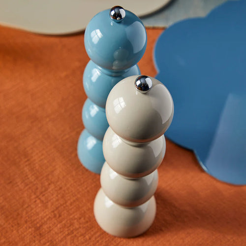 Stacked sphere design salt and pepper grinder set. One is light blue and the other is beige. Silver cap on top. Sitting on an orange colored surface with a blue and white plate on the side. 
