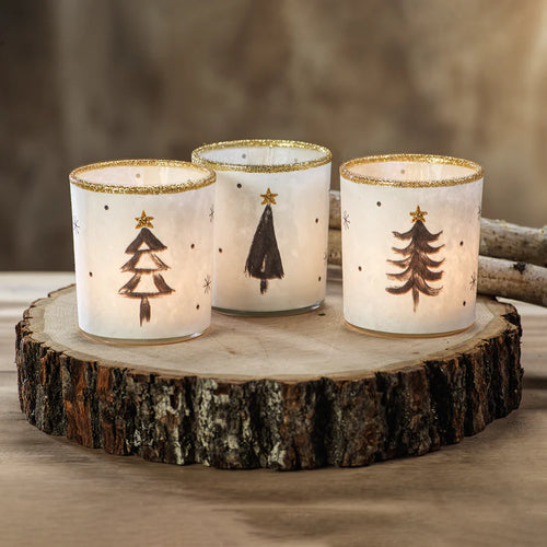 Three white tea light holders with gold glitter rims and black Christmas tree designs, each glowing with a lit candle, displayed on a wood slice surface against a neutral blurred background. 