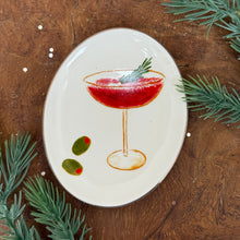 Load image into Gallery viewer, White , oval,  ceramic dish with a cocktail glass and  a red drink design . Green fir tree cutting as a garnish. Two green olives at the bottom of the dish. On a wooden surface, surrounded by fir tree cuttings.