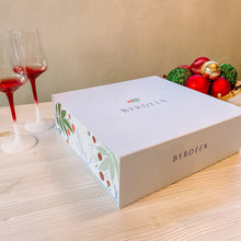 Load image into Gallery viewer, Closed Brydeen gift box with a white lid and festive floral patterned sides displayed on a light wooden surface beside two red-to-white gradient wine glasses and a cluster of red, green, and gold holiday ornaments.