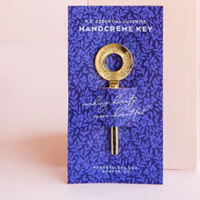 Load image into Gallery viewer, A gold colored hand creme key on blue packaging against a pink surface.