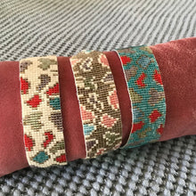 Load image into Gallery viewer, Three different beaded banned bracelets wrapped around a red cushion. First bracelet is cream with brown, red, and blue leopard spots. Second bracelet is brown with pink, blue, and brown leopard spots. The third bracelet is teal with red, blue, and brown leopard spots. Sitting on top of a gray textured surface.