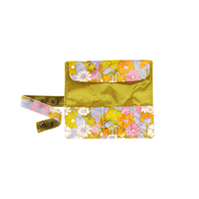 Load image into Gallery viewer, Floral makeup brush roll with a mustard yellow interior and a pink, orange, lavender, and white retro flower pattern, shown opened and empty with matching snap-closure straps, displayed against a black and gray checkered background.   