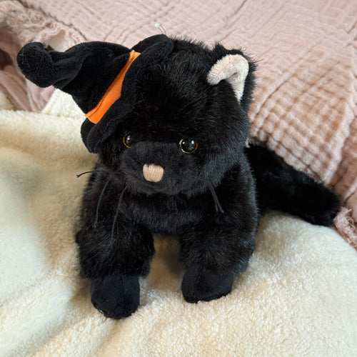 Black plush cat wearing a black witch hat with an orange band, with a tan nose, brown eyes, black whiskers, and white ears on a white plush surface with a pink textured textile in the background. 