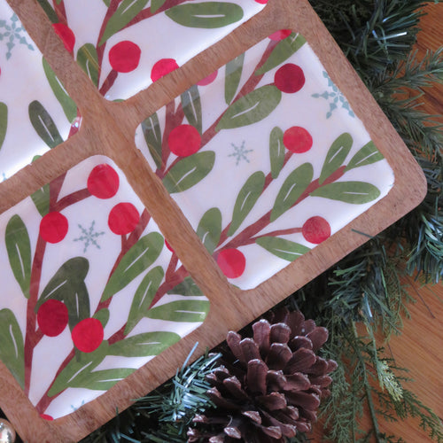 Close-up of a four-section mango wood and white ceramic tray featuring a winter berry design with green leaves, red berries, and light blue snowflakes, displayed on a wooden surface with a pinecone and green garland.  