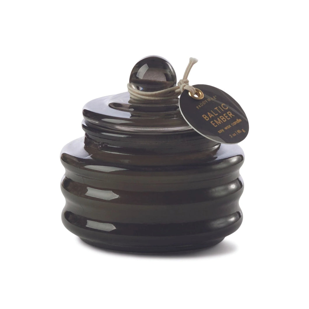 Dark grey glass candle with a visible brand tag on a white background. Body of the jar is ribbed shaped withs knob on top. Tag has the text 