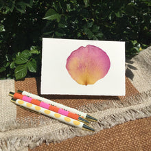 Load image into Gallery viewer, Greeting card with a pressed pink flower petal with various colored ink pens on a beige  and white blanket and bishes in the background. 
