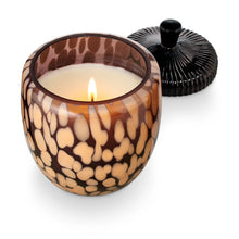 Load image into Gallery viewer, Midnight Pumpkin Glass Acorn Candle, view from above of the open glass jar with irregular tan spots and a dark brown background filled with cream-colored wax and a single lit wick, with its glossy black ridged lid resting behind it against a white background.