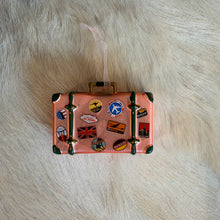 Load image into Gallery viewer, Traveler Luggage Ornament, lifestyle overhead view of a pink painted glass suitcase ornament with dark green straps and corner accents, gold-colored buckles and handle, colorful travel-sticker illustrations, and a sheer white hanging ribbon, resting on a cream fur-textured surface.