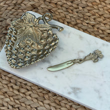 Load image into Gallery viewer, White Marble Cheese Tray, close-up of a white marble serving tray with subtle gray veining, a gold-tone textured strawberry-shaped bowl with a large veined leaf and curled stem, and a matching spreader with a twisty vine handle and leafy end on a brown woven fiber surface. 