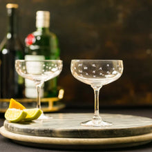 Load image into Gallery viewer, Two stemmed glasses with small star etchings on stacked round cutting boards. Two lime slices and a lemon slice are on the board. Two wine bottles are behind the display. A black and amber wall is the background. 