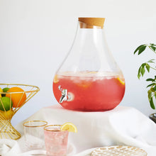 Load image into Gallery viewer, Clear glass beverage dispenser with a silver-colored spigot and a cork stopper, filled with a red drink and citrus slices, styled with drinkware, greenery, and a gold-colored fruit basket filled with an orange, lemons, and limes, on a white cloth-covered surface. 