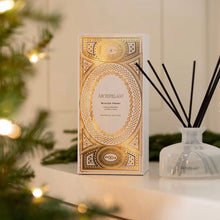 Load image into Gallery viewer, Archipelago Winter Frost glass diffuser with black reeds next to its metallic gold and white packaging box on a white surface with a lit blurred Christmas tree in the foreground.