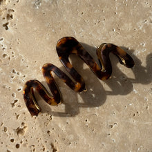 Load image into Gallery viewer, Tortoiseshell wavy claw clip with gold metal hardware displayed on a beige travertine surface with soft shadows. 