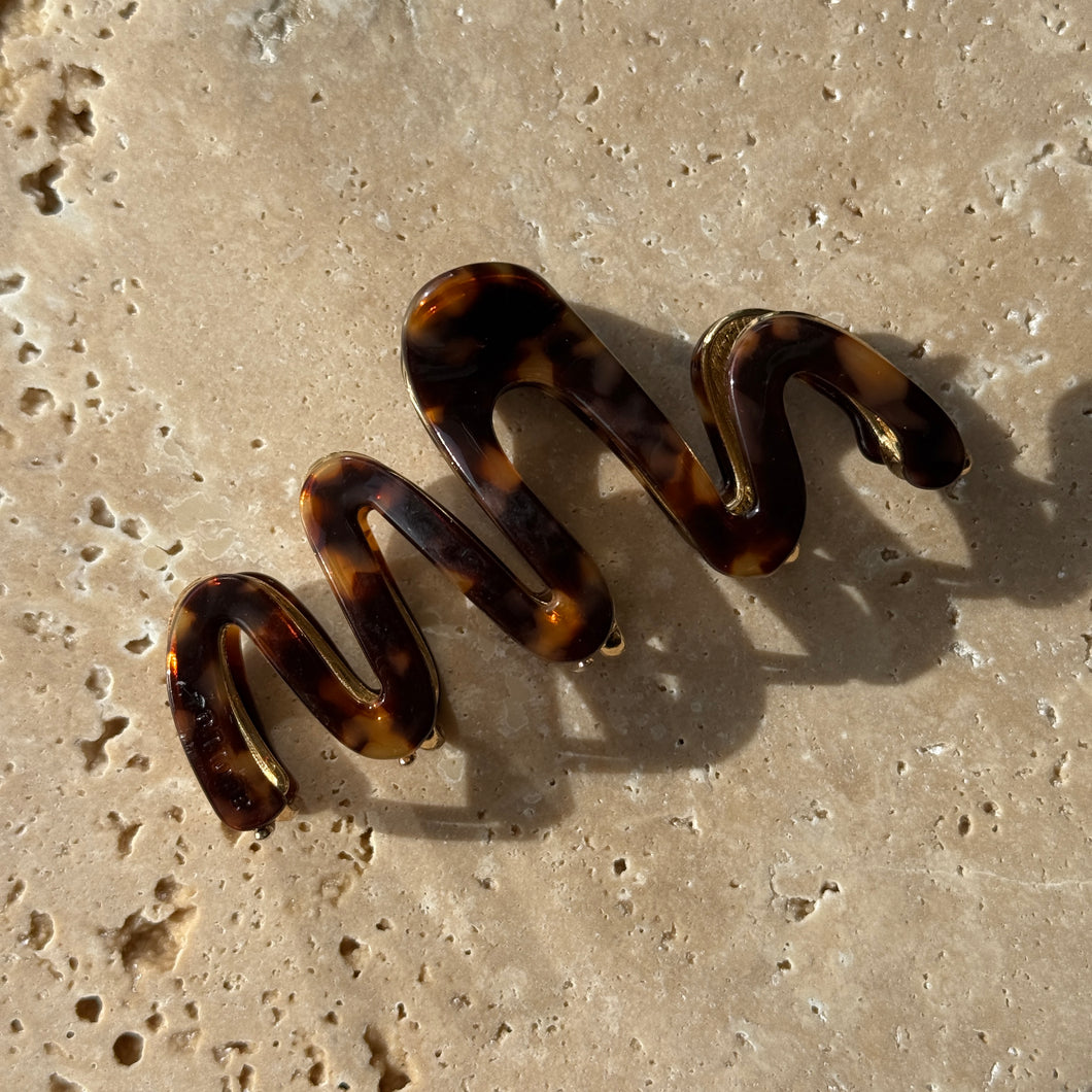 Tortoiseshell wavy claw clip with gold metal hardware displayed on a beige travertine surface with soft shadows. 