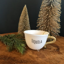 Load image into Gallery viewer, White teacup ornament with a gold glitter handle , a clear footed bottom, and &quot;TEQUILA&quot; in black text. Two decorative frosted Christmas trees , and a green fir tree cutting. On a wooden surface , against a dark background.