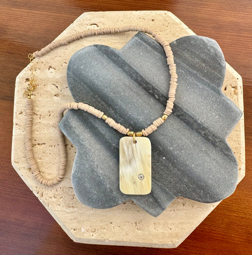 Necklace featuring a polished rectangular stone pendant with a single clear round accent stone, strung on neutral disc beads with small gold accents, displayed on a quatrefoil-shaped gray stone tile layered over a beige travertine slab on a wooden surface. 