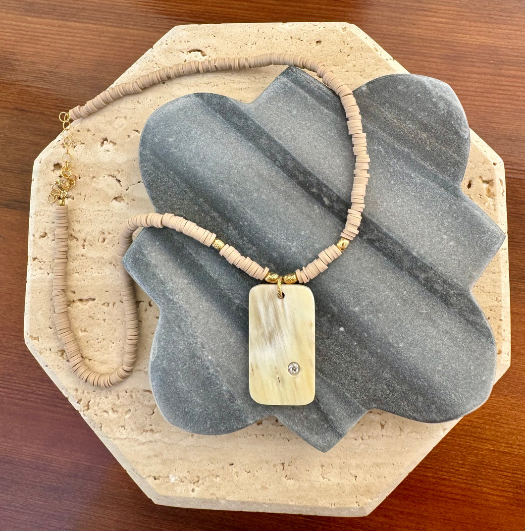 Necklace featuring a polished rectangular stone pendant with a single clear round accent stone, strung on neutral disc beads with small gold accents, displayed on a quatrefoil-shaped gray stone tile layered over a beige travertine slab on a wooden surface. 