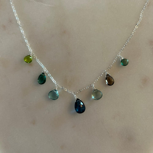 Silver necklace with 7 multicolored teardrop gem charms in various shades of blue and green. White marble background. 