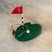 Load image into Gallery viewer, Putting Green Ornament, lifestyle angled view of a painted glass putting green with a glittery green base, black golf hole, small white golf ball, and red-and-white striped flagpole topped with a red “Best Christmas By Par” flag, with a silver-colored hanging cord, resting on a cream fur-textured surface.
