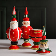 Load image into Gallery viewer, Group of three hand-painted ceramic cookie jars featuring Santa Claus, Mrs. Claus, and a nutcracker in red outfits with  gold star details, and tall finials in red, green, and white. Displayed on a white surface with the nutcracker jar open and its lid placed in front.