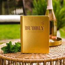 Load image into Gallery viewer, Gold cocktail recipe book "BUBBLY" by Colleen Jeffers styled outdoors with a bottle and fresh mint.