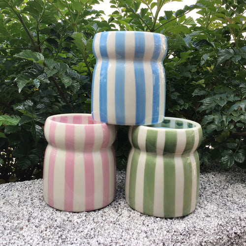 Three ceramic striped candles stacked in pyramid shape. Pink and green ceramic striped  candles on the bottom, and a blue striped ceramic candle on top. Sitting on a  stone surface with a green bush in the background. 