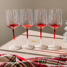 Load image into Gallery viewer, Set of four hand-blown crystal wine glasses featuring red-to-white gradient stems and bases, arranged in a row on a sunlit white wooden surface with a red plaid blanket in the foreground. 