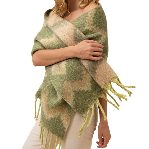Person standing against a white background, draped in an oversized reversible knit scarf featuring an olive, beige, and cream zigzag and diamond pattern with light green fringe. 