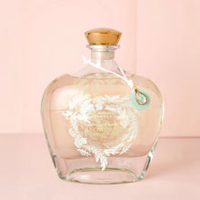 Load image into Gallery viewer, Clear glass Snowdrop bubble bath bottle with frosted detailing, gold accents, a cork stopper, and a decorative crystal charm displayed against a powder pink background. 