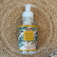 Load image into Gallery viewer, Balsam &amp; Fir, lifestyle image showing an overhead view of foaming hand wash in a clear, rounded plastic bottle with a white pump and a beige wraparound label featuring green evergreen branches, brown pinecones, and a gold-colored panel with the scent name and product type in dark lettering, resting on a brown woven mat.